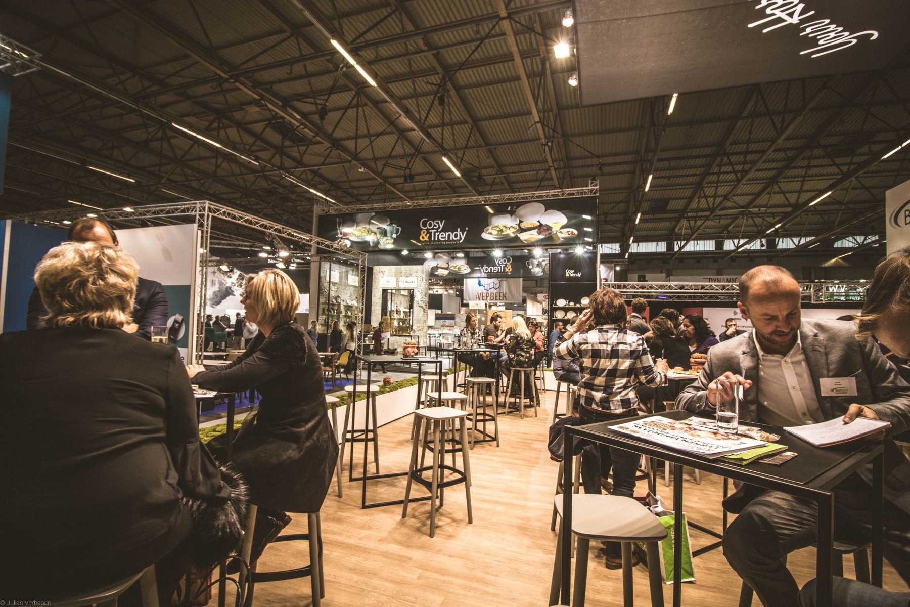 Wide trade-show environment showing a completed booth, visitors, furniture and branded exhibition structures working together.